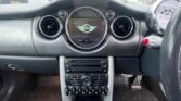 The interior dashboard of a MINI car showcases the central control panel, with the iconic MINI logo displayed on the middle screen. The dashboard features circular air vents, various control buttons, and a manual gear stick in the foreground. The speedometer is visible on the right side.