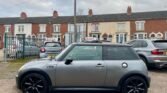 A silver MINI Cooper with black wheels is parked on a gravel area in front of a row of attached brick houses. The sky is overcast, and there are two other cars nearby: one directly behind and the other to the right. Utility wires stretch across the cloudy sky above the scene.