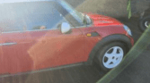 A red mini car with a white roof is parked on a paved surface in bright sunlight. The sun creates glare and visible lens flares across the image. A brick wall and part of another vehicle are visible in the background.