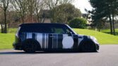 A black, low-profile MINI with large black rims and custom striped camouflage paint featuring vertical gray, white, and black lines and a large white circle on the side is parked on a paved surface. It’s set against a backdrop of green grass, leafless trees, and a partially visible house.