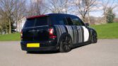 A customized black MINI Clubman car is parked on a paved road near a grassy area with leafless trees. The MINI features a unique striped and circular design in shades of gray, black, and white on its rear, sides, and roof. The rear windows are heavily tinted, and the license plate is yellow.