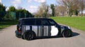A black Mini Clubman car is parked on a paved road beside a grassy area with bare trees. This unique mini car is covered in monochrome camouflage with vertical stripes and a large white circle under a partly cloudy sky.