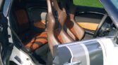 The image shows the interior of a small MINI car with the driver's side door open. The vehicle features sporty brown and black bucket seats, and the upholstery has a mix of leather and suede sections. The two-door design allows a clear view of the stylish passenger seat as well.