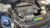 A fully opened Mini Cooper hood displays the engine compartment, showing the engine block with the "MINI" logo, air filter, coolant reservoir, and various hoses and wires. The car's circular headlight and front grille are also visible in the foreground.