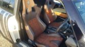 The interior of the MINI features two brown and black leather racing seats with angular headrest openings and perforated patterns. The door on the left is open, revealing the seats more clearly, while the driver's side shows a steering wheel and gear shift. Sunlight casts shadows inside.