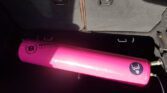 A pink racing gas canister, labeled "BAKER Motorsport," is secured in the trunk of a MINI vehicle. The canister also features a black logo of a stylized animal head. The surrounding interior is dark, with two folding seat latches visible in the background.