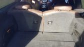 View of the backseat and trunk area of a MINI with brown leather seats and a black interior. The backseats are upright, dividing the trunk space. Various controls and dials are visible on the car's dashboard through the gap between the seats. The trunk area appears spacious and carpeted.