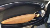 The image showcases the interior door panel of a MINI, featuring a sleek, curved design in black material. The door handle is distinctively shaped like a longboard or surfboard, covered in tan, suede-like material, adding a unique and stylish touch to the car's interior.