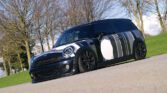 A low-profile black MINI Clubman with white and gray abstract stripes is parked on a paved surface. It features a modified front bumper, blacked-out wheels, and tinted windows. The background consists of leafless trees and a green grassy area under a blue sky with scattered clouds.