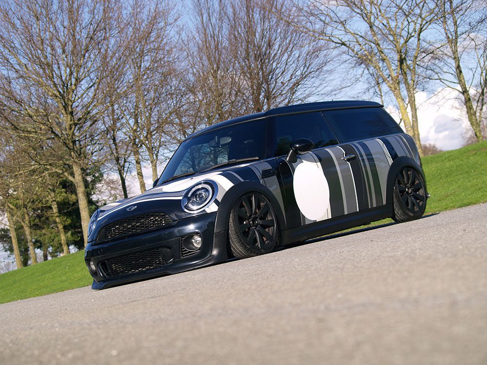 A low-profile black MINI Clubman with white and gray abstract stripes is parked on a paved surface. It features a modified front bumper, blacked-out wheels, and tinted windows. The background consists of leafless trees and a green grassy area under a blue sky with scattered clouds.