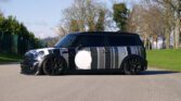 A black MINI Clubman with a sleek, custom wrap featuring vertical grey and white stripes is parked on a paved road. The car's body sits low to the ground, its glossy finish reflecting the surrounding greenery and trees. Bright blue sky with scattered clouds can be seen in the background.