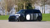 A modified black MINI Cooper Clubman with a striped camouflage paint job is parked on a paved surface. The car has a custom lower suspension, black wheels, and a white circular decal on the driver's door. Leafless trees and grassy areas are visible in the background under a clear sky.