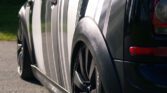 Close-up of the rear side of a black MINI with white and grey vertical stripes. The vehicle has sleek, shiny black wheels and well-defined body curves, reflecting sunlight, and is parked on a paved surface. The background shows a blurry green area, indicating an outdoor setting.