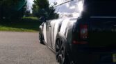 A black MINI Clubman car with a prominent white stripe pattern is parked on a paved road. The car’s glossy surface reflects sunlight, creating a bright glare on its rear window. The scene includes a grassy area and some trees in the background under a partly cloudy sky.