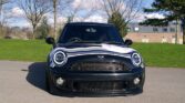 A black MINI Cooper is parked on a paved surface in front of a grassy area with leafless trees and a building in the background. The car's distinctive circular headlights and rounded hood are prominent. The sky is partly cloudy with patches of blue.