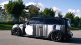 A matte gray MINI Cooper Clubman with black and white vertical stripes painted on the sides. The car features black tinted windows, black wheels, and modified lowered suspension. It is parked on an asphalt surface beside a lawn with bushes, trees, and a metallic wire fence in the background.
