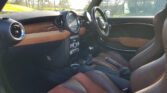 Interior view of a modern MINI car with brown leather seats and black trim. The dashboard features a large central speedometer, with additional dials and controls below. The steering wheel is on the right side, indicating a right-hand drive vehicle. Sunlight enters through the windows, illuminating the interior.