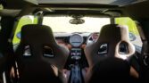 Interior view of a compact MINI from the back seat, showcasing black bucket seats with cutouts, a central dashboard with a large speedometer, various control knobs, and a sporty steering wheel. The car's sunroof is partially visible at the top, allowing sunlight to enter.