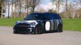 A customized black MINI Cooper Clubman with unique white and gray geometric patterns is parked on a paved surface. The background features leafless trees and green grass under a partly cloudy sky.