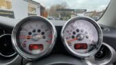 Close-up of a MINI car dashboard featuring a speedometer and tachometer. The speedometer shows 98 mph, while the tachometer is at roughly 3,000 RPM. The digital odometer displays 84,175 miles. The background shows a cloudy sky and a gas station.