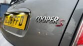 Close-up of the rear badge on a grey MINI Cooper S with "COOPER S" written in chrome lettering on the right side of the hatchback. Below the car's rear window, there is a UK license plate with the number "GL06 FFD" in black characters on a yellow background.