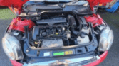 A red mini car with its hood open reveals the engine bay and various components. Parked on a paved surface, the engine parts, battery, and fluid reservoirs are clearly visible beneath the raised hood.