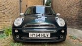 A black MINI Cooper with the license plate "HY54 HLV" is parked in a narrow passageway between two brick walls. The car faces forward, its headlights illuminated. The ground is paved with worn bricks, and there are some weeds growing in the cracks. A building with a window is visible in the background.
