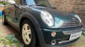 A dark green MINI Cooper with a white roof and mirrors is parked in an outdoor, cobblestone area surrounded by brick walls. The car has a license plate reading "HY54 AWO" and displays distinctive round headlights and a chrome front grille. There are small plants growing between the cobblestones.