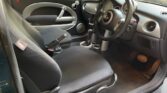 The interior of this MINI compact car features a driver's seat, steering wheel, and dashboard. The seats are upholstered in dark fabric, while the door panels boast contrasting light and dark sections. The dashboard is equipped with multiple dials and controls, complemented by a gear stick between the front seats.