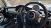 Interior of a MINI car showcasing the dashboard and steering wheel from the driver's seat perspective. The dashboard features several gauges, including a central speedometer, and the steering wheel has a central emblem. The gear shift and cup holders are also visible in the foreground with greenery outside.