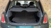 A green MINI hatchback car is pictured with its trunk door open, revealing the empty cargo space. The car is parked on a brick driveway, and the interior is mostly obscured by the trunk cover. The rear seats and back of the driver's seat are partially visible through the open trunk.