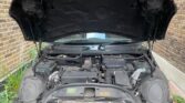The image shows an open MINI car hood revealing the engine compartment. The car is parked next to a brick wall. The engine parts, including various cables and components, are visible but slightly dusty. There are some plants growing next to the car, and part of a building can be seen in the background.