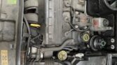 A photo of an engine bay from a Mini car, showcasing various components like the air filter housing and coolant reservoir. The engine cover prominently displays the brand "MINI" in yellow lettering, surrounded by typical mechanical details and connectors.