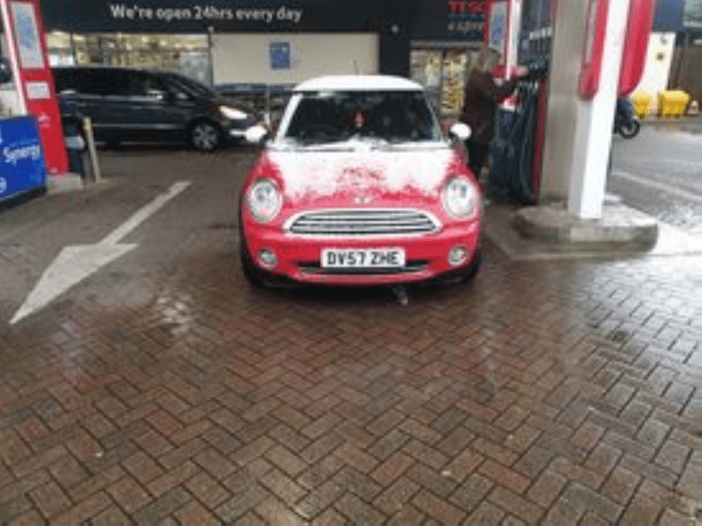 A red mini car with a white roof, dusted with snow, is parked at a gas station. The car faces forward at the pump. The ground is wet, and a black van is in the background. A sign above reads, "We're open 24 hrs every day.