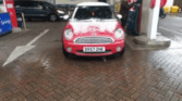A red mini car with a white roof, dusted with snow, is parked at a gas station. The car faces forward at the pump. The ground is wet, and a black van is in the background. A sign above reads, "We're open 24 hrs every day.