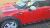 A red Mini Cooper car with a white roof, white side mirror, and white wheels is parked on a sunny street. The iconic mini car’s driver-side door and front wheel are visible, with tree shadows falling across the windshield and hood.