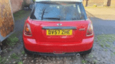 A red mini car with a white roof is parked on a cobblestone area. The car’s rear license plate reads "DV57 ZHE." There are buildings, a shed, and a bicycle visible in the background on a sunny day.