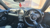 View of a mini car’s interior from the back seat, showing a black dashboard with a round central speedometer, navigation display, circular air vents, manual gear stick, and steering wheel with Mini logo. Sunlight pours through the front windshield.