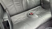 A mini car’s back seat upholstered in black leather with curved stitching patterns. Two headrests are visible, and the seatbelt buckles are centered. The interior features dark carpeting and cup holders in the lower right corner.