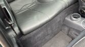 The image shows the rear seat of a mini car with dark leather upholstery. The center has a raised, rounded section with two red and black seat belt buckles. The floor is carpeted, and there are cup holders and a storage net on the front seat back.