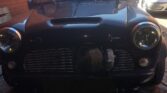 A dark blue classic Mini car is parked on a brick driveway between red brick buildings. The Mini has round headlights, a chrome grille, and a prominent turbocharger visible through the lower grille area. Another car is parked in the background—perfect for any Mini Trader enthusiast.