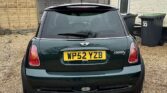A dark green mini car with license plate WP52 YZB is parked on a gravel driveway in front of a cream brick house with a brown roof. Two black wheelie bins labeled 55 stand on the right, and a wooden fence is on the left—a true find for any Mini trader.