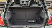 A green mini car with its rear hatch open reveals the empty trunk and folded seats. The car's interior, dashboard, and steering wheel are visible through the back. This Mini is parked on a gravel driveway near a house, ready for any mini trader adventure.