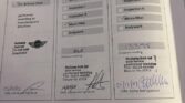 A service booklet for a mini car displays three maintenance entries. Each is stamped, signed, and dated, with boxes checked for Oil Service, Inspection I/II, Micro-filter, and Bodywork. Handwritten mileages: 4415 and 12398; booklet rests on a wooden desk.