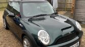 A dark green Mini car with silver alloy wheels is parked on a gravel driveway. The car has a UK license plate (WP52 YZB). Behind it, a wooden fence, shed, laundry on a clothesline, leafless trees, and two red-brick houses appear—a true mini trader scene.