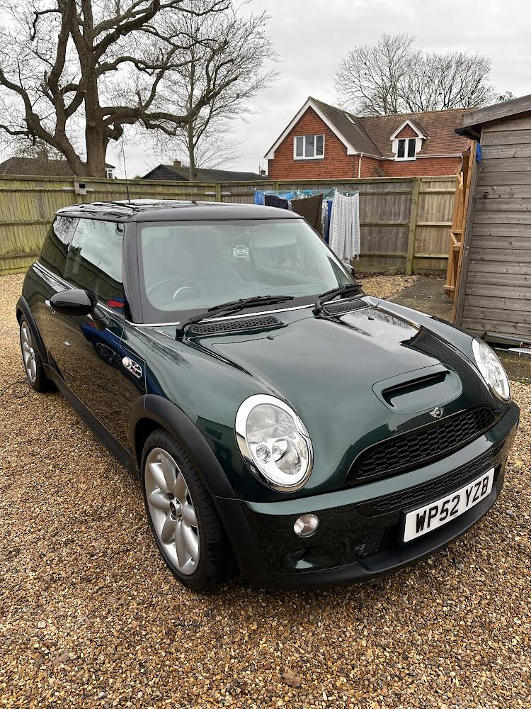 A dark green Mini car with silver alloy wheels is parked on a gravel driveway. The car has a UK license plate (WP52 YZB). Behind it, a wooden fence, shed, laundry on a clothesline, leafless trees, and two red-brick houses appear—a true mini trader scene.