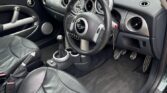 The image shows the black leather interior of a right-hand drive Mini Cooper S, a classic mini car. The steering wheel, dashboard with circular gauges, manual gear shift, and contoured seats with some creasing are visible.