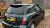 A dark green Mini Cooper S with a black roof and silver rims is parked on a gravel driveway. This classic mini car, with UK plate "WP52 YZB," sits in front of wooden fencing, outbuildings, and part of a house with a tiled roof.