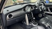 A photo of a Mini car’s interior taken from the passenger side, showing black leather seats, a manual gear stick, a large round speedometer in the center dashboard, and a right-hand steering wheel. The doors and dashboard are gray and silver.