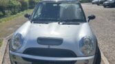 A silver Mini Cooper from a mini trader is parked at an angle in a sunny outdoor lot, front wheels turned right. The car’s UK license plate reads “SG54 XFY.” Trees and other parked cars are visible under a partly cloudy sky.